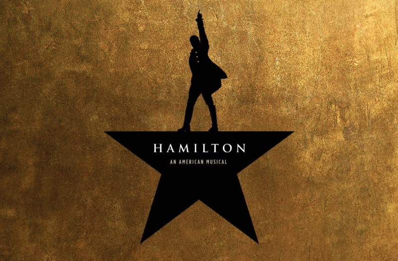 The banner image reads Hamilton An American Musical, 11 Tony Awards, The Pulitzer Prize. Against a textured gold background, a man in 18th century frock coat and top boots stands in silhouette, turned slightly sideways, his left arm pointing upwards.