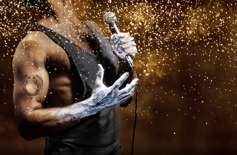 The image for Jesus Christ Superstar is a close-up photograph of the torso of a muscular black man, turned sideways to us. His neck is tensed upwards and he holds a microphone in his left hand, his right hand fingers outstretched. Both of his hands up to halfway up the forearm are covered in silver paint or make-up and he has a double-angel tattoo on his bicep. Gold sparkles fill the air.