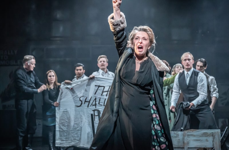 The image for Merchant of Venice is a production photo. Close up to the front is Tracy-Ann Oberman as Shylock. She raises her fist defiantly. Behind her other characters in shirtsleeves hold a home-made banner which reads: They Shall Not Pass.