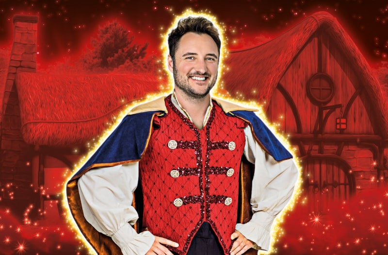 James Bye as Prince Charming stands resplendent in doublet and cape, hands on his hips, in front of a red backdrop of a rustic thatched cottage. A glittery gold halo surrounds him.