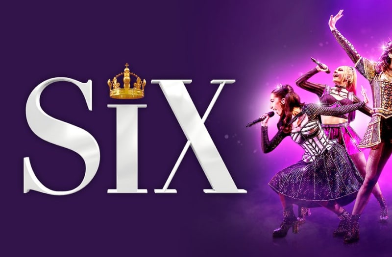 The banner image for Six has the title in capital letters to the left, with a gold crown standing in for the dot of the i. To the right the six queens hold mics, singing and throwing shapes, dressed in their colourful, radically-updated mediaeval costumes.