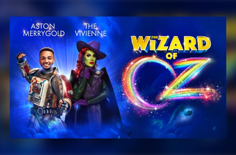 Aston as a robot-like Tin Man stands bravely to the left of this image, with The Vivienne to his left, posing quizzically as the Wicked Witch. She wears a purple fedora hat and a black cape. To the right is The Wizard Of in block gold letters and Oz in sparkly rainbow colours.