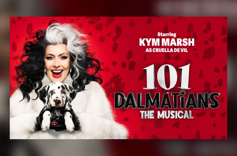 Kym Marsh as Cruella smiles with white teeth and bright red lips, her wild black hair streaked with white. She wears a white fur coat and holds a little dalmatian puppy tightly in her grasp.