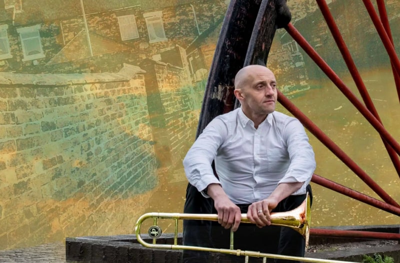 A photographic montage shows Shane, a middle-aged shaven-headed man wearing a shirt, holding his trombone. In the background is a pit-head winding wheel and a nostalgic view of the backyards of terraced houses.