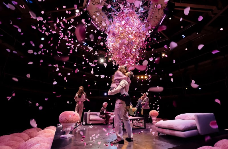 A stylish production photo shows the characters in modern dress on a glossy black square stage surrounded by fluffy pink bean bags. Confetti fills the air as Gwendolen leaps into Jack’s arms, in the background Algernon and Cecily and Dr Chasuble and Miss Prism embrace. Lady Bracknell sits alone on a low sofa.