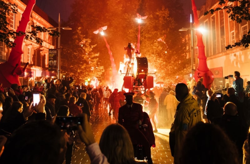 Photograph shows an atmospheric street scene at night, glowing winged creatures on long stalks soar above the audiences’ heads as a procession passes down the middle of the street.