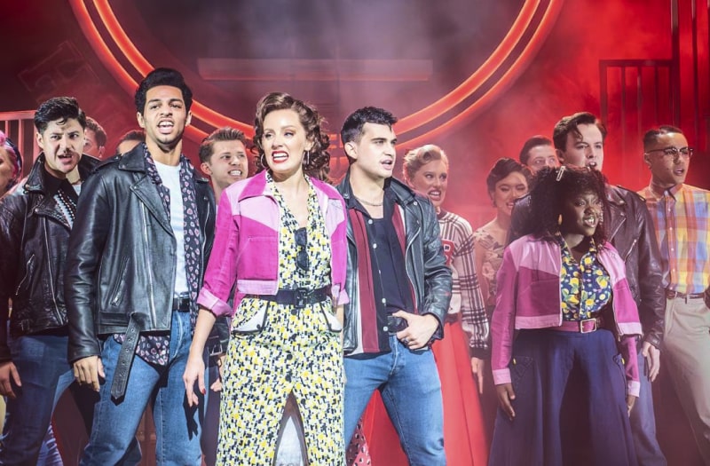 A production photo shows the full cast onstage mid musical number. They are a mixture of young men, Danny and his gang in black motorcycle jackets and jeans, others in chinos and casual shirts. The women mostly wear 50s style casual clothes, Frenchie’s gang with pink western-style jackets.