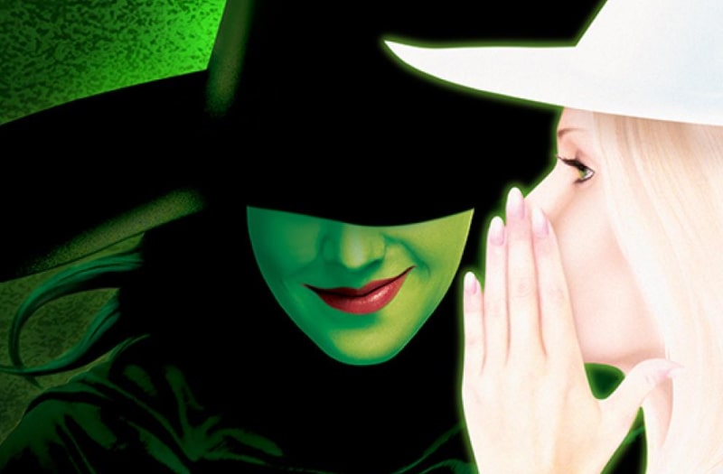 The image has the Wicked Witch on the left, dressed in black, her green face almost obscured by the brim of her hat. Her red lips are twisted in a smirk. To the right, dressed in white and in profile, Glinda whispers a secret into her ear.