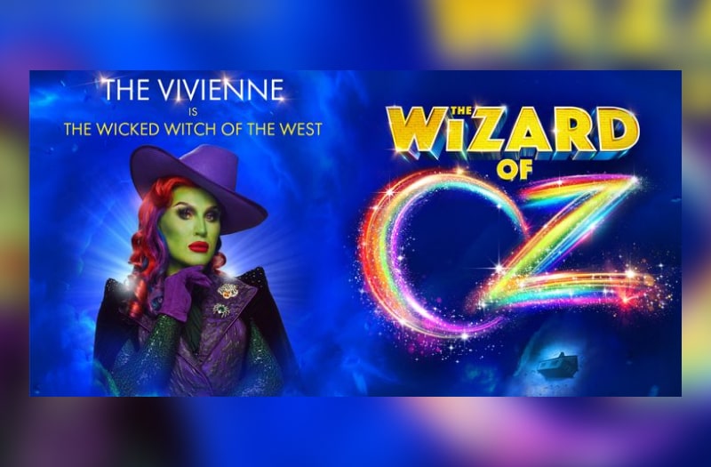 The Vivienne with stylish fedora hat, red locks and black cape poses quizzically as the Wicked Witch, chin on gloved hand. To the right is The Wizard Of in block gold letters and Oz in sparkly rainbow colours and above her reads The Vivienne The Wicked Witch of the West.
