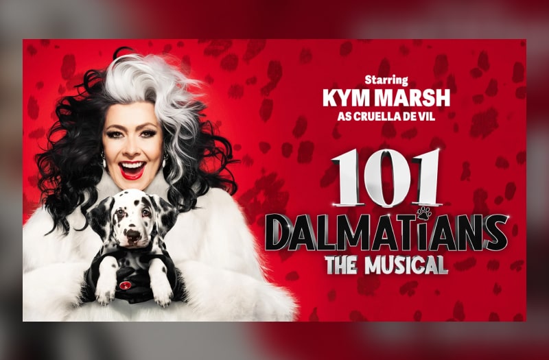 Kym Marsh as Cruella smiles with white teeth and bright red lips, her wild black hair streaked with white. She wears a white fur coat and holds a little dalmatian puppy tightly in her grasp.