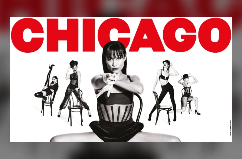 The image has the show title in red capitals. Below it five women are photographed in black and white in provocative postures using chairs as props. They each wear sexy fetish lingerie. The woman in the centre is close up, sitting astride her chair, pointing her finger at us as though it were a gun.