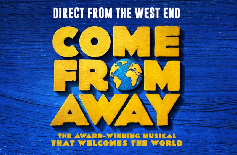 The banner image has Come From Away in block capitals in a pale brown colour as though made of wood. The O of From is a tiny drawing of the Earth. The background is a deep blue, textured as though of thick paint.