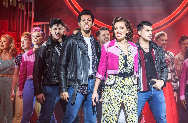 A production photo shows the full cast onstage mid musical number. They are a mixture of young men, Danny and his gang in black motorcycle jackets and jeans, others in chinos and casual shirts. The women mostly wear 50s style casual clothes, Frenchie’s gang with pink western-style jackets.