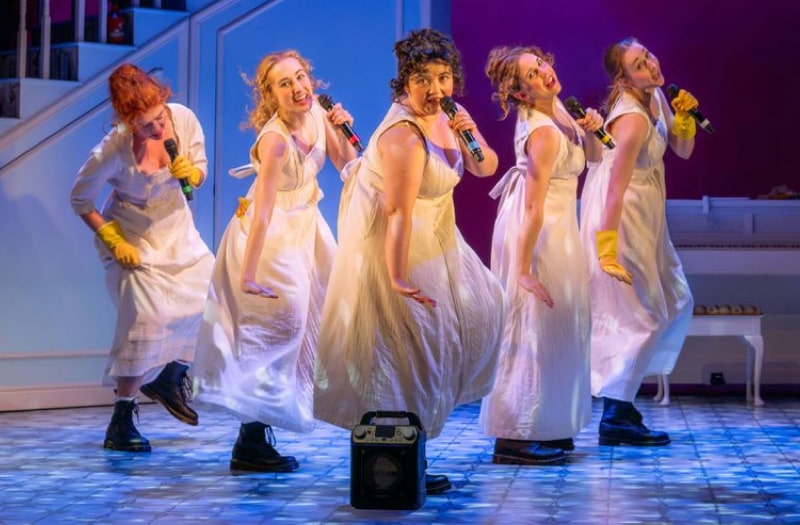 Five young women stand in a v shape, dancing and sing karaoke as they hold microphones. They are dressed in high-waisted white muslin gowns and black Doc Marten boots. At the back of the stage set is a white piano.