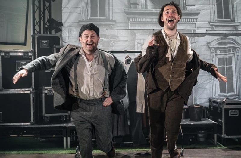 A production photo shows Charlie and Jake onstage against a theatre backdrop of a Georgian house and piles of flight cases. Charlie is shorter and well filled out. He wears a crumpled grey suit with braces and a flat cap turned backwards. Jake on the right is taller and wears a brown suit and brown moleskin waistcoat, his cap also turned the wrong way round. They seem to be running, with joyous expressions on their faces.